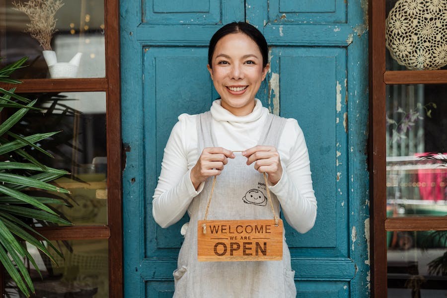 A woman standing outside of her business in front of a rustic blue door, holding an “open” sign. 