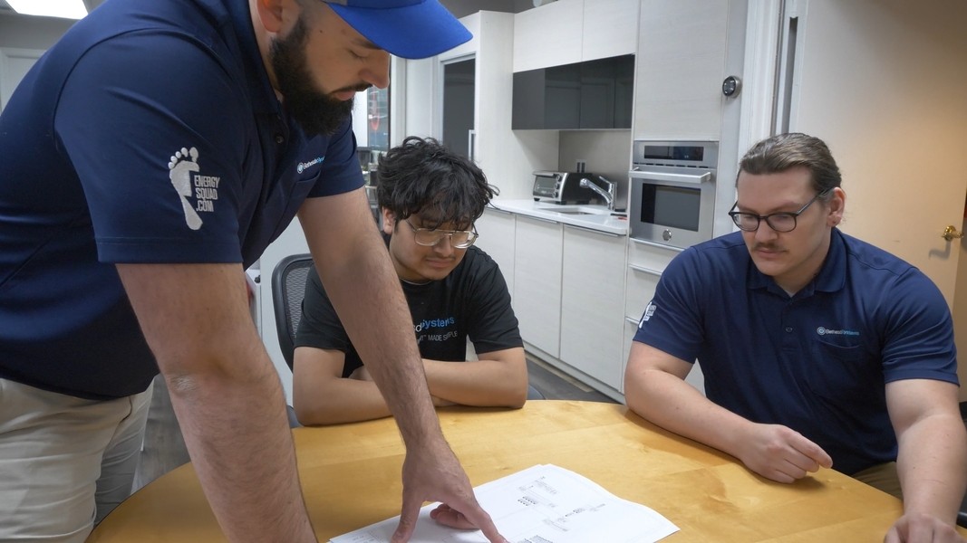 Three Bethesda Systems employees sit at a round table in a modern kitchen-style office as one of them points to system documentation on the table.